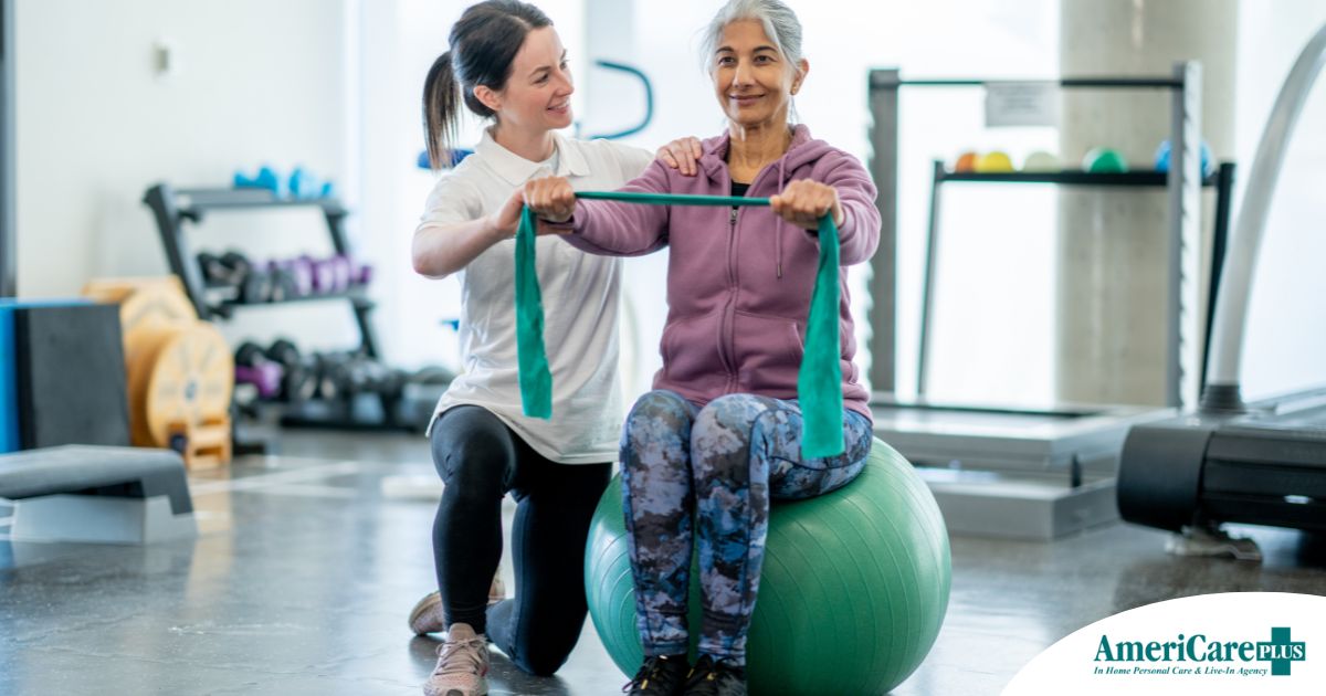 A care provider helps an older woman exercise with a resistance band and an exercise ball, representing how exercise can help with senior fall prevention.
