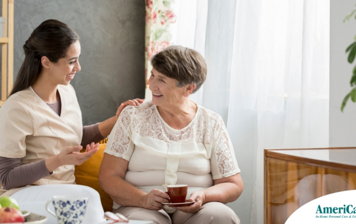 Smiling caregiver with her hand on an older woman’s shoulder as they share tea on the couch, showing home care vs assisted living