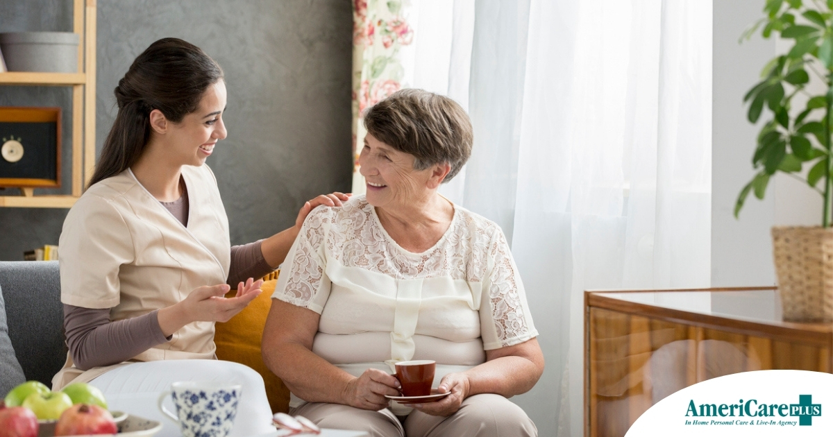 Home Care vs Assisted Living Where Caregiving Work Fits in Today’s Families Smiling caregiver with her hand on an older woman’s shoulder as they share tea on the couch, showing home care vs assisted living
